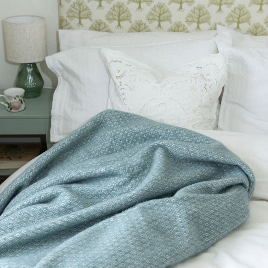 Harald dusty blue wool throw on a bed with white bed linen and a green bedside table