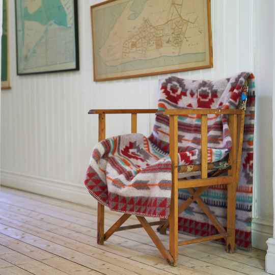 Wooden chair with a patterned blanket draped over it, against a white wall with framed maps.