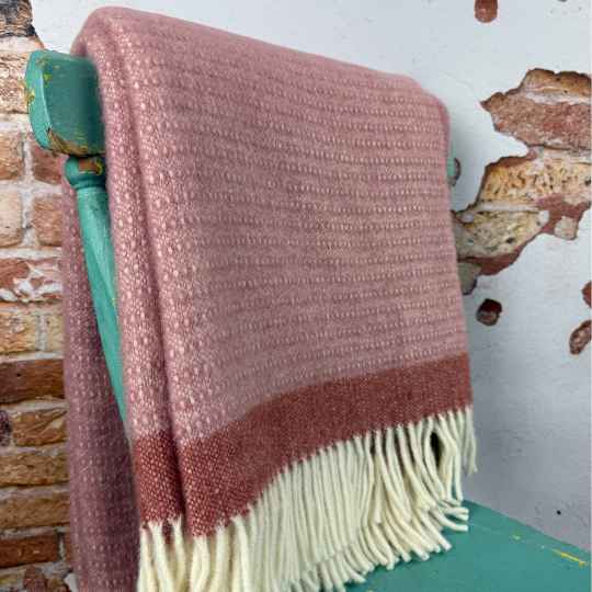 Pink textured throw blanket with fringes draped over a green chair against a rustic brick wall.