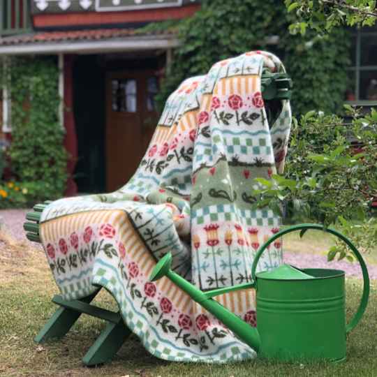 Floral-patterned blanket draped over a green chair in a garden setting with a watering can nearby.