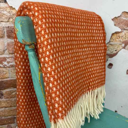 Orange woven wool blanket with white fringe draped over a teal chair against a brick wall background