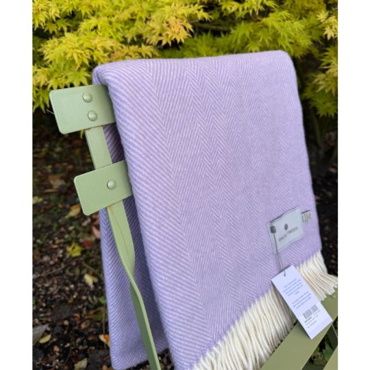A lilac and cream herringbone patterned throw blanket draped over a light green metal garden chair, with a white product tag attached.