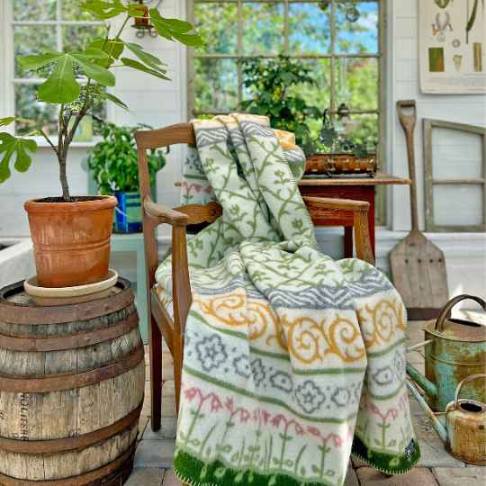 Decorative blanket with a floral pattern draped over a chair in a sunlit room with plants and a barrel.