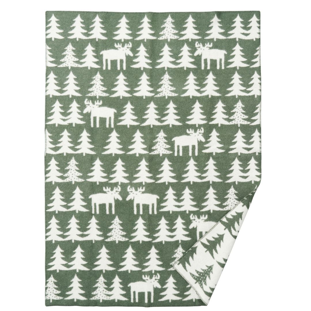 Wool blanket l, featuring a repeating pattern of white moose and white Christmas trees on a muted sage green background. The bottom right corner is folded over to show the reverse side, which has a white background with a green tree pattern.