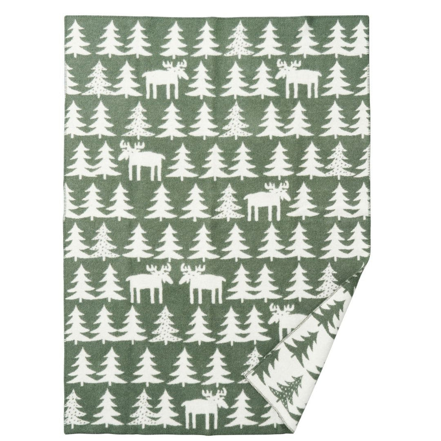 Wool blanket l, featuring a repeating pattern of white moose and white Christmas trees on a muted sage green background. The bottom right corner is folded over to show the reverse side, which has a white background with a green tree pattern.