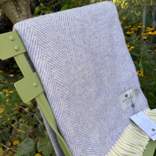 A light lavender and cream herringbone woven throw blanket draped over a light green folding chair in an outdoor garden setting.
