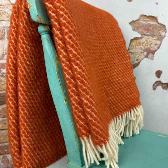 Orange wool blanket with white fringe draped over a teal chair against a textured wall.
