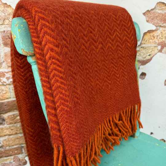 Orange wool blanket with fringes and a zig zag pattern draped over a turquoise chair against a wall.
